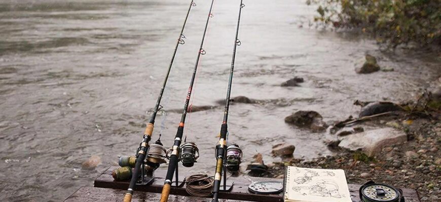 Рыболов на рассвете у реки выбирает фидер: несколько удилищ с разными вершинами и кормушками на влажной скамье, мягкий пасмурный свет.