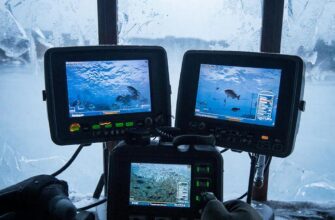 Рыбак в ледовой палатке сравнивает несколько зимних эхолотов на скамье: теплый свет внутри, синяя стужа и метель за окном, пар дыхания и сияющие экраны.