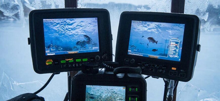 Рыбак в ледовой палатке сравнивает несколько зимних эхолотов на скамье: теплый свет внутри, синяя стужа и метель за окном, пар дыхания и сияющие экраны.