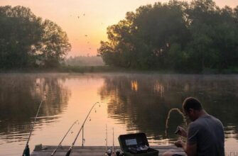 Рыбак готовит приманку из манной крупы на берегу тихого озера на рассвете, в туманной атмосфере и мягких красках восходящего солнца.